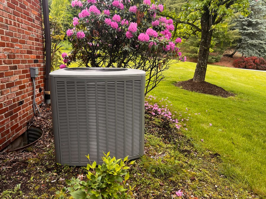 Air conditioner unit outside of a house during Spring. Pink flowers are in the garden beside it.