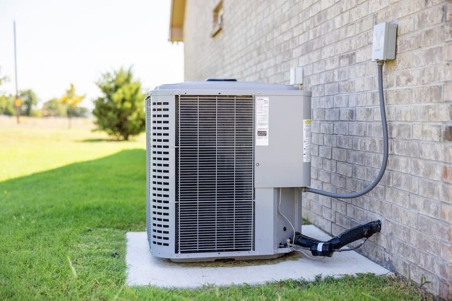 Outdoor air conditioning unit on the side of a brick house with grass and trees in the background